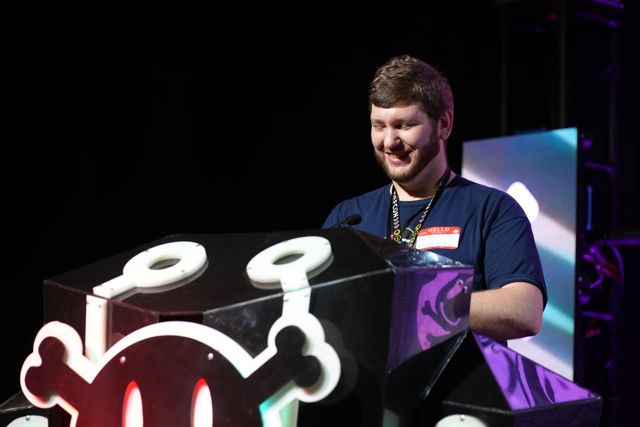 A person standing behind a podium with a cartoonish skull and crossbones design, smiling and wearing a dark blue shirt with a name tag