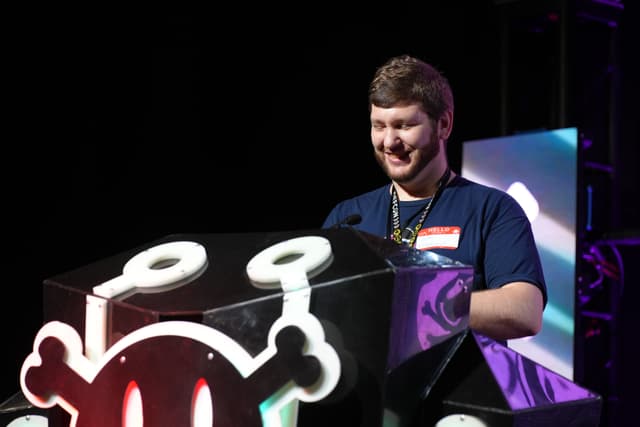 A person standing behind a podium with a cartoonish skull and crossbones design, smiling and wearing a dark blue shirt with a name tag