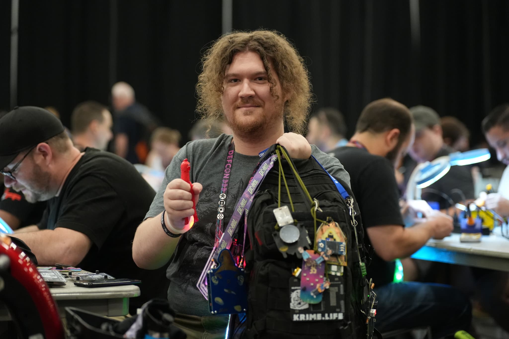 A person with curly hair and a beard, wearing a lanyard with various badges and holding a red object, stands in a room filled with people working at tables