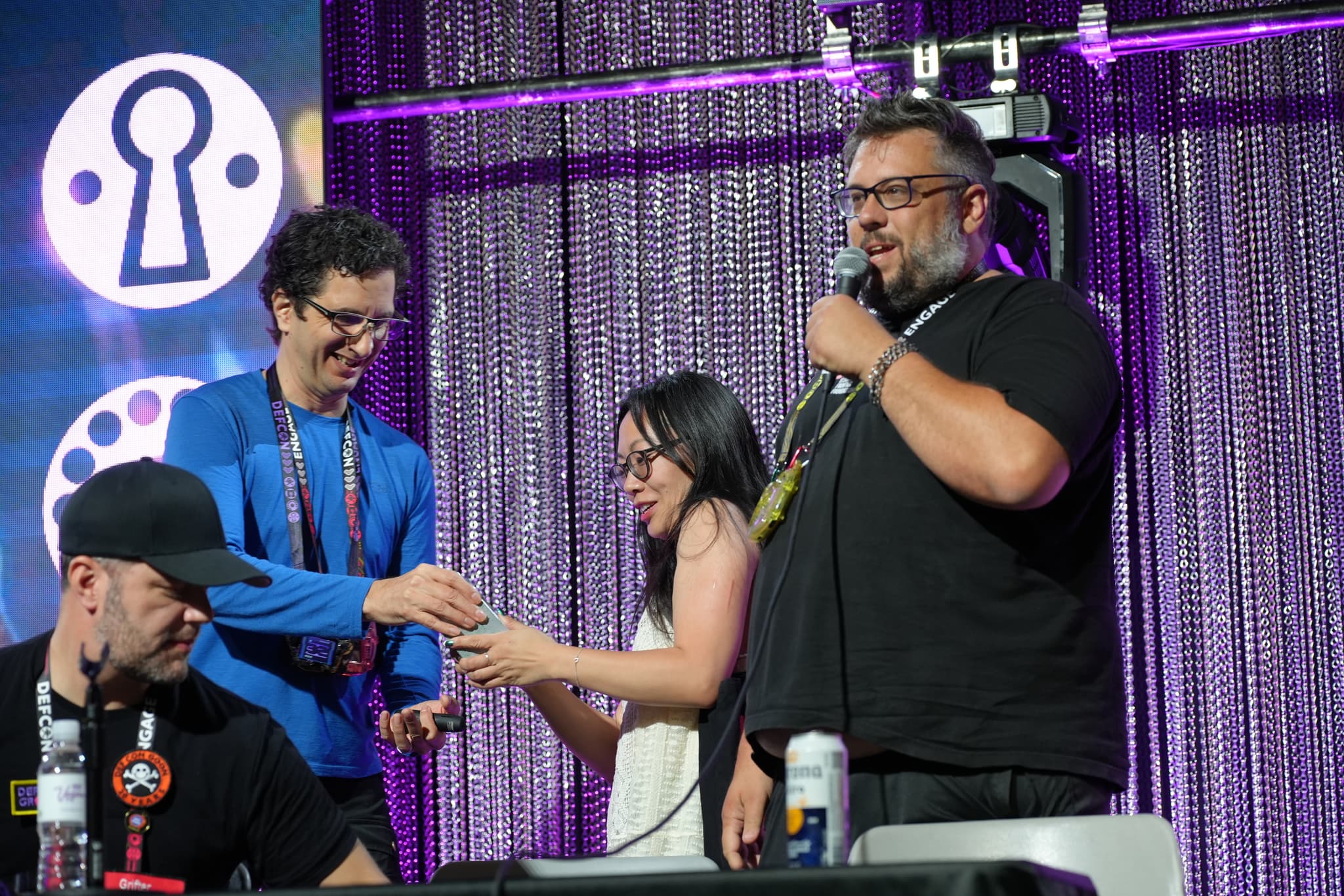 Four people on a stage, with one person speaking into a microphone. Two individuals are exchanging an item, while another person is seated and looking down. The background features a purple curtain and a logo with a keyhole design