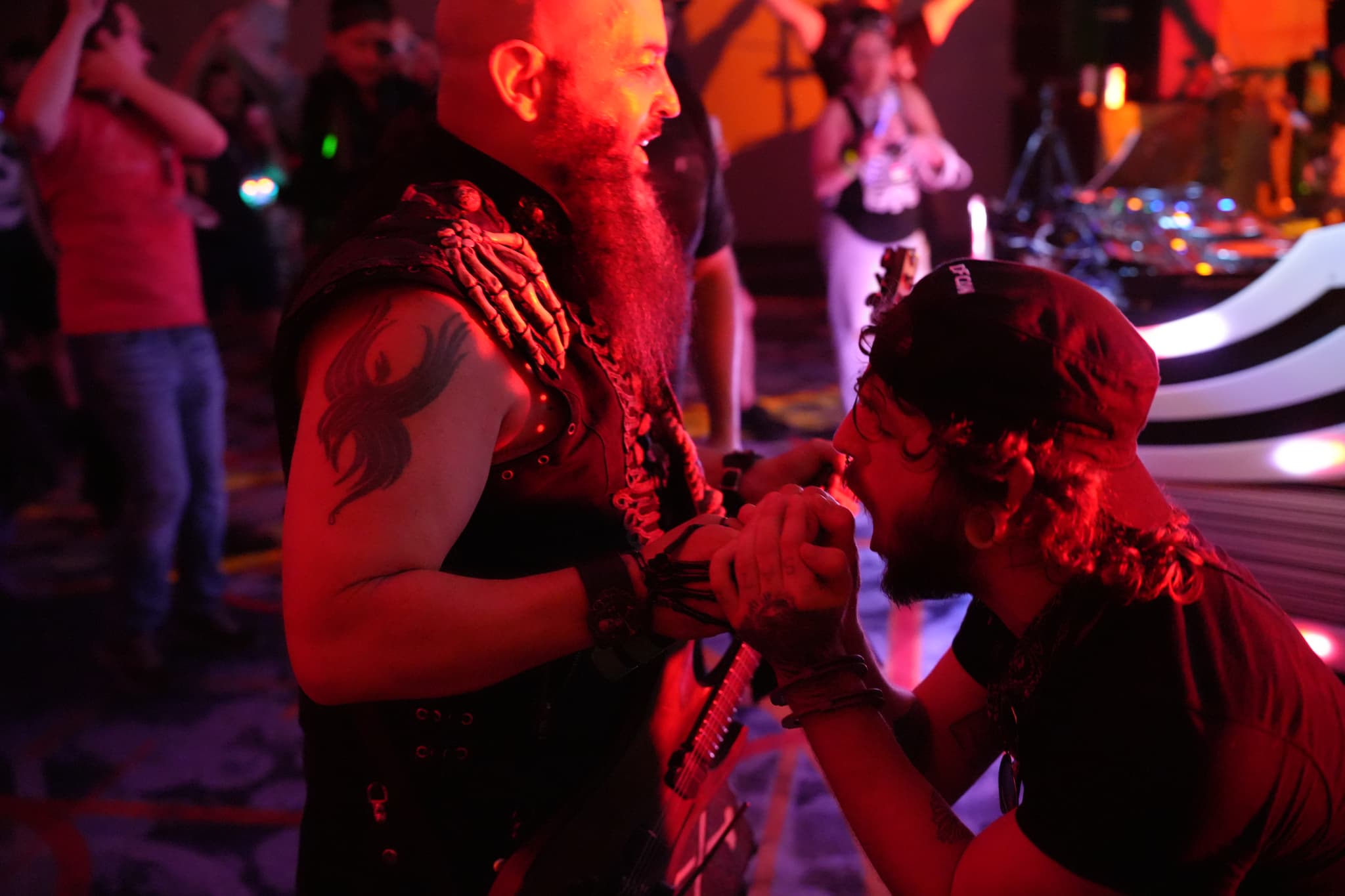 Two men in a dimly lit room, one with a beard and tattoo, the other kneeling and holding a microphone, surrounded by people and colorful lights