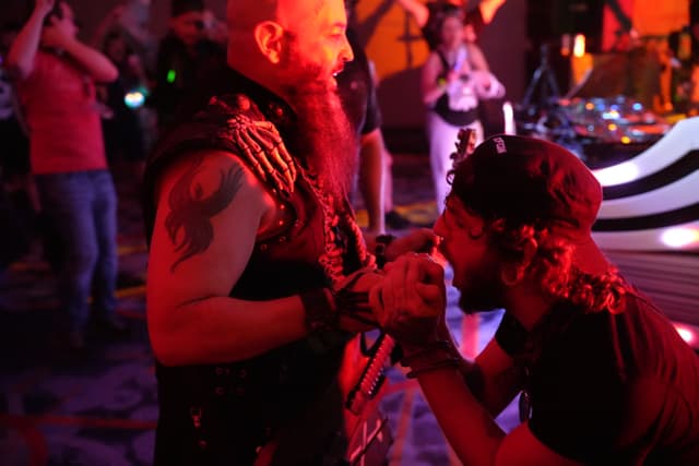 Two men in a dimly lit room, one with a beard and tattoo, the other kneeling and holding a microphone, surrounded by people and colorful lights