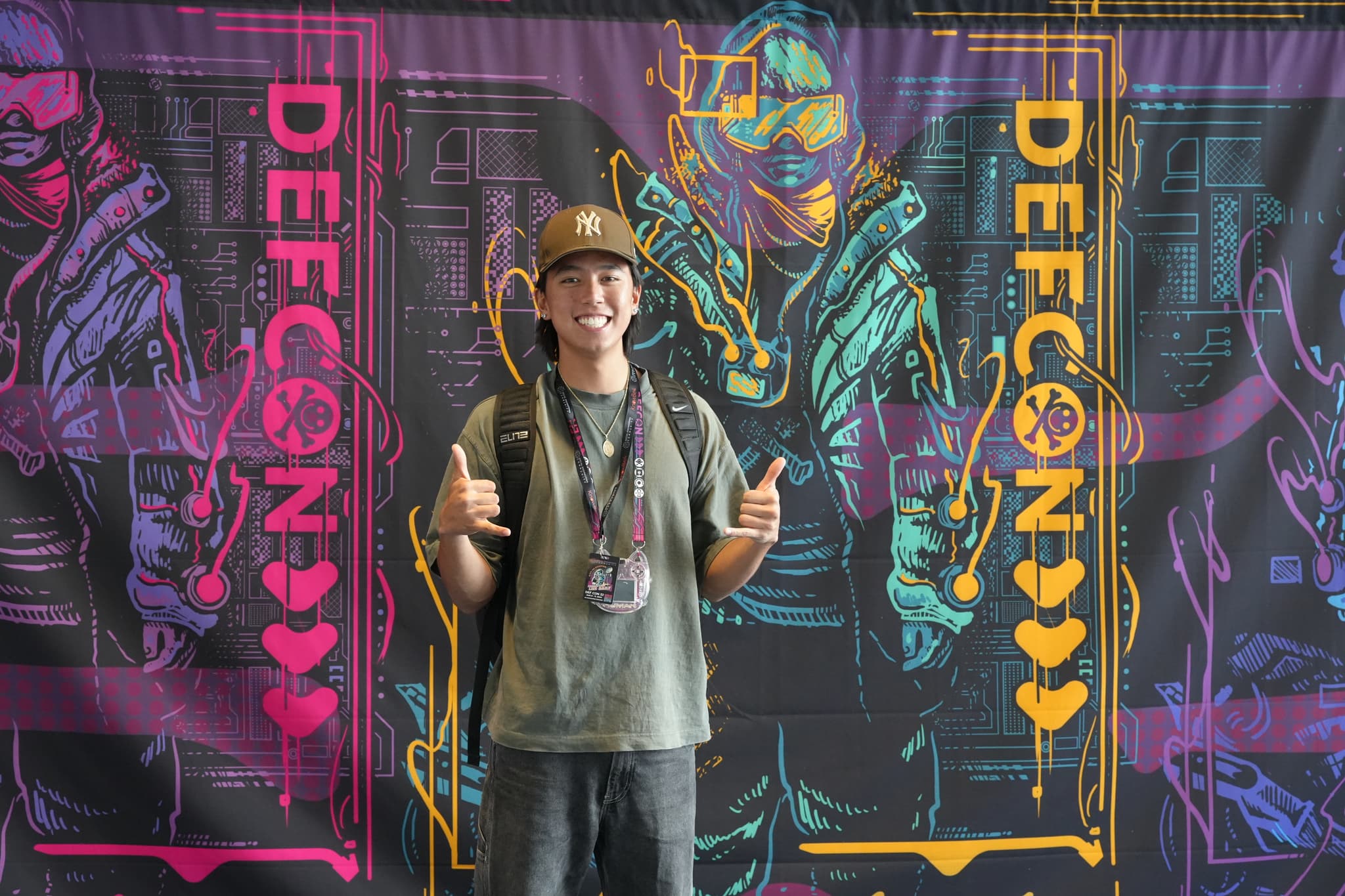 A person wearing a hat and backpack stands in front of a colorful Defcon-themed backdrop, giving a thumbs-up gesture with both hands