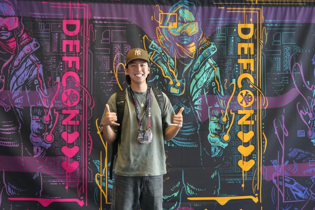 A person wearing a hat and backpack stands in front of a colorful Defcon-themed backdrop, giving a thumbs-up gesture with both hands