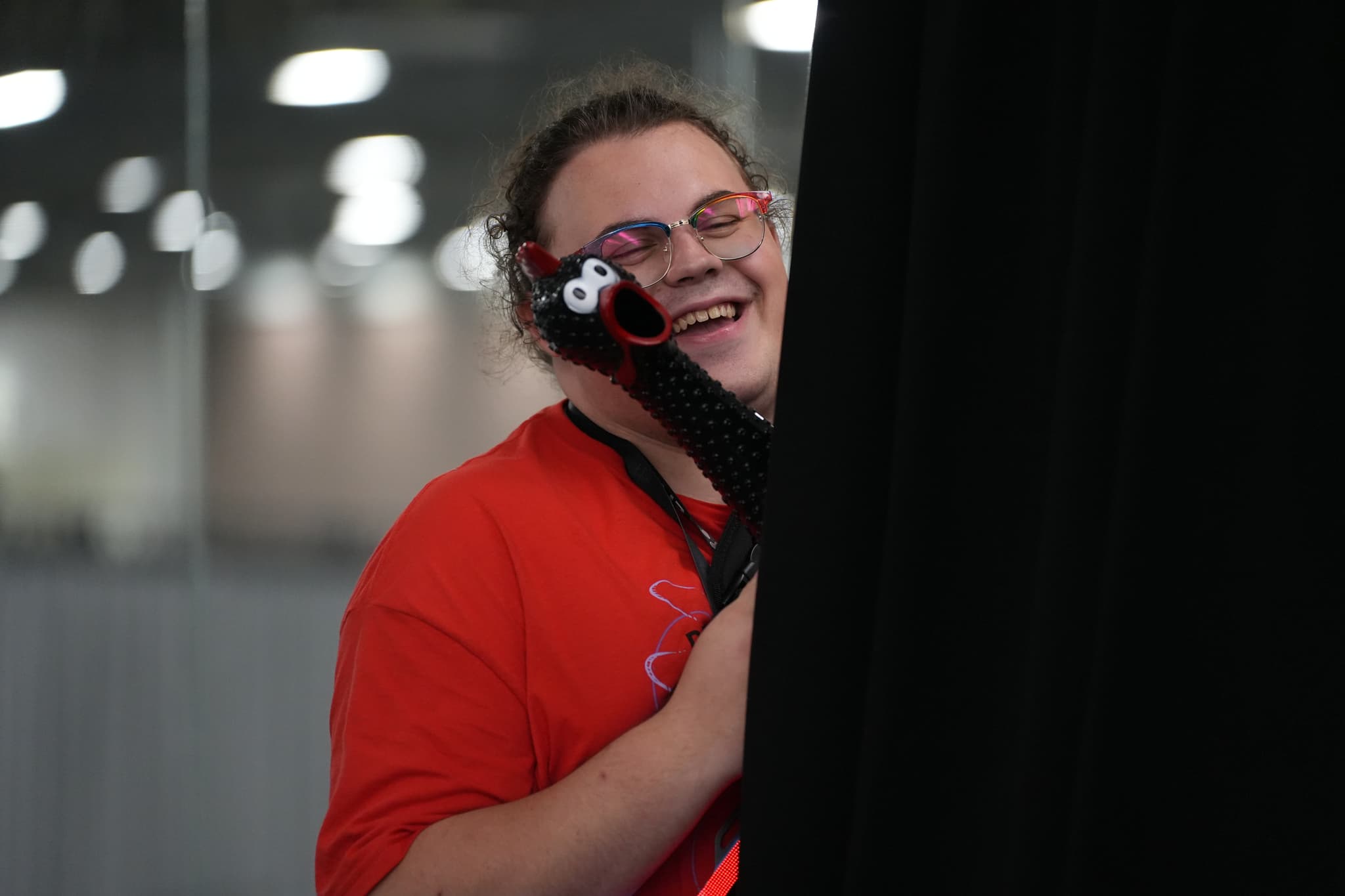 A person wearing a red shirt and glasses is smiling while holding a black object, with a blurred background of lights and structures