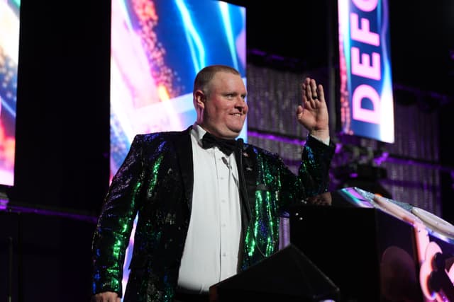 A man in a shiny, sequined jacket and bow tie is standing on a stage, raising his hand in a wave. The background features colorful lights and large screens with text