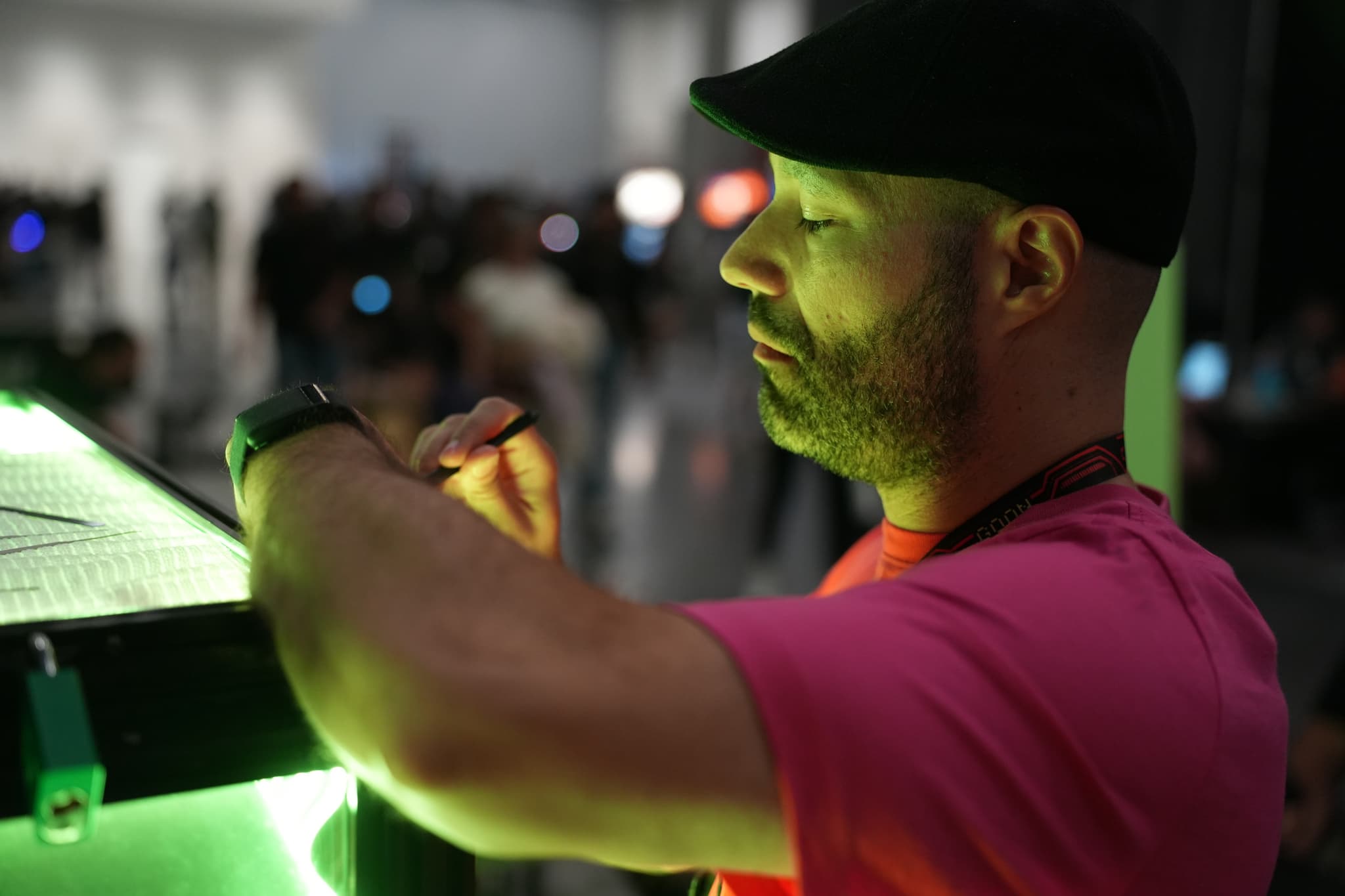 A man in a pink shirt and black cap is interacting with a green-lit device in a dimly lit environment with blurred people in the background