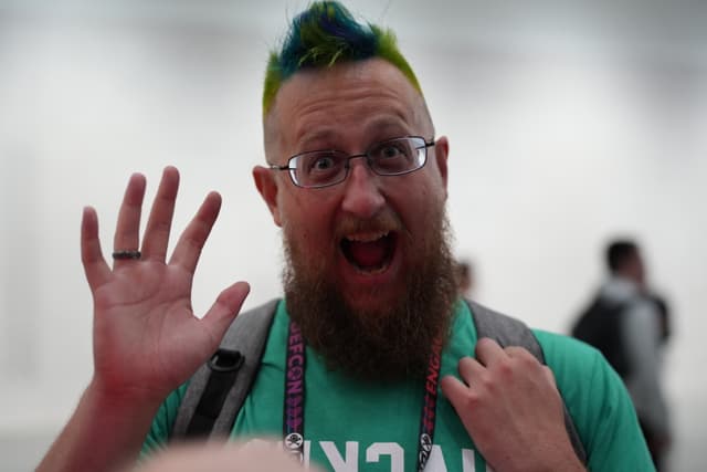 A bearded person with glasses and a colorful mohawk hairstyle is enthusiastically waving with a wide-open mouth, wearing a green shirt and a backpack