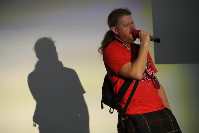 A person with long hair, wearing a red shirt and a kilt, is speaking into a microphone. A shadow of the person is cast on the wall behind them