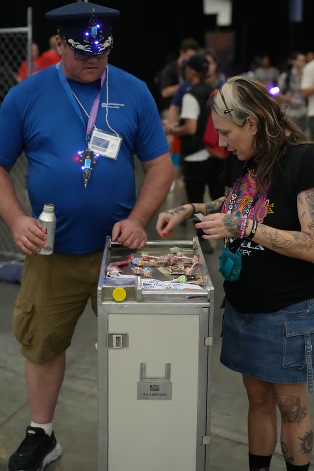 Two people are standing next to a small cart with various items on it. The person on the left is wearing a blue shirt, khaki shorts, and a blue hat, holding a drink. The person on the right is wearing a black shirt and a denim skirt, looking at the items on the cart. There are other people and a fence in the background