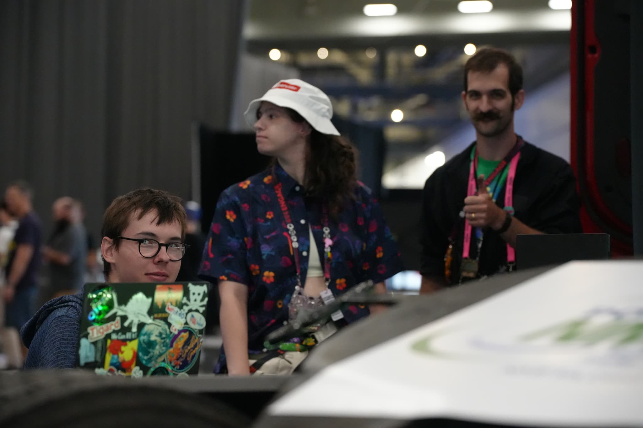 Three people are gathered around a table with laptops, one person is wearing a white hat, and another is holding a drink. The background appears to be an indoor event or conference