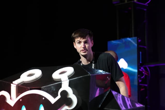 A person standing behind a podium with a stylized design, possibly at an event or performance, with stage lighting and equipment in the background