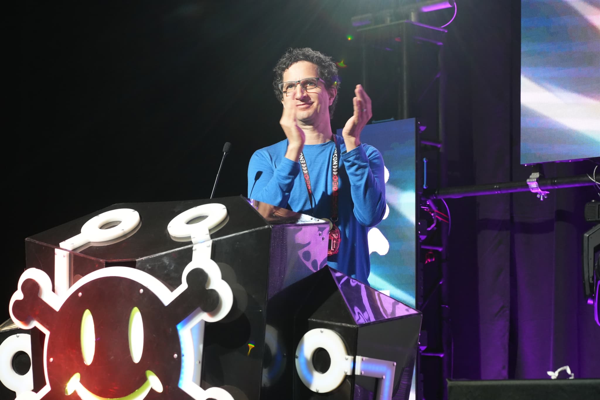 A person standing behind a podium decorated with a stylized skull and crossbones, clapping their hands, with stage lighting and equipment in the background