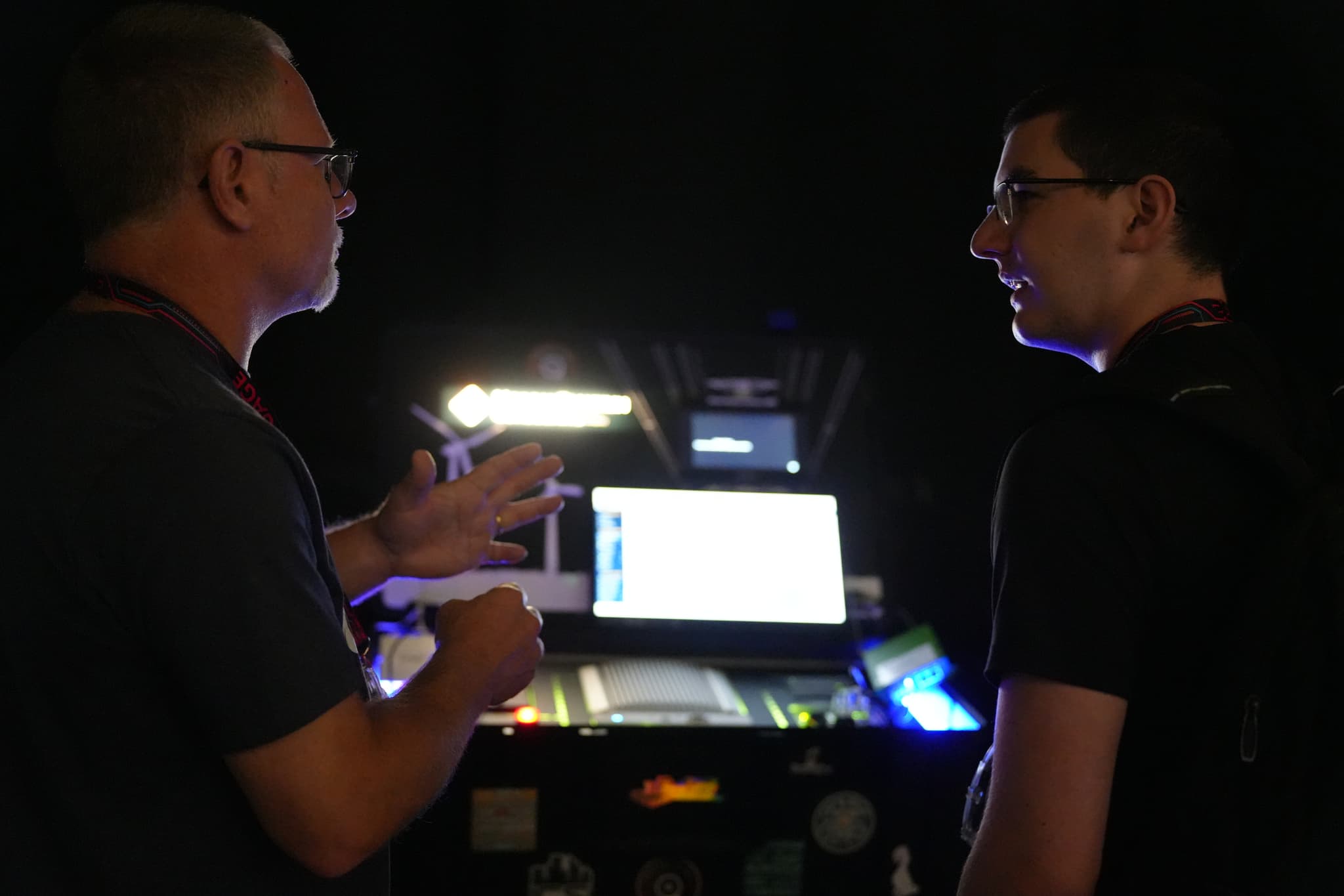 Two people engaged in conversation in a dimly lit room, with a computer and various equipment in the background