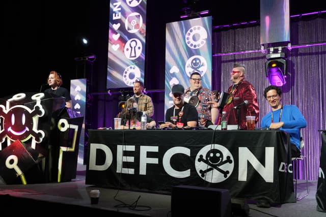 A group of people on a stage at DEFCON, with a large banner displaying the DEFCON logo. The individuals are standing or sitting behind a table, and the background features various tech-themed decorations and lighting