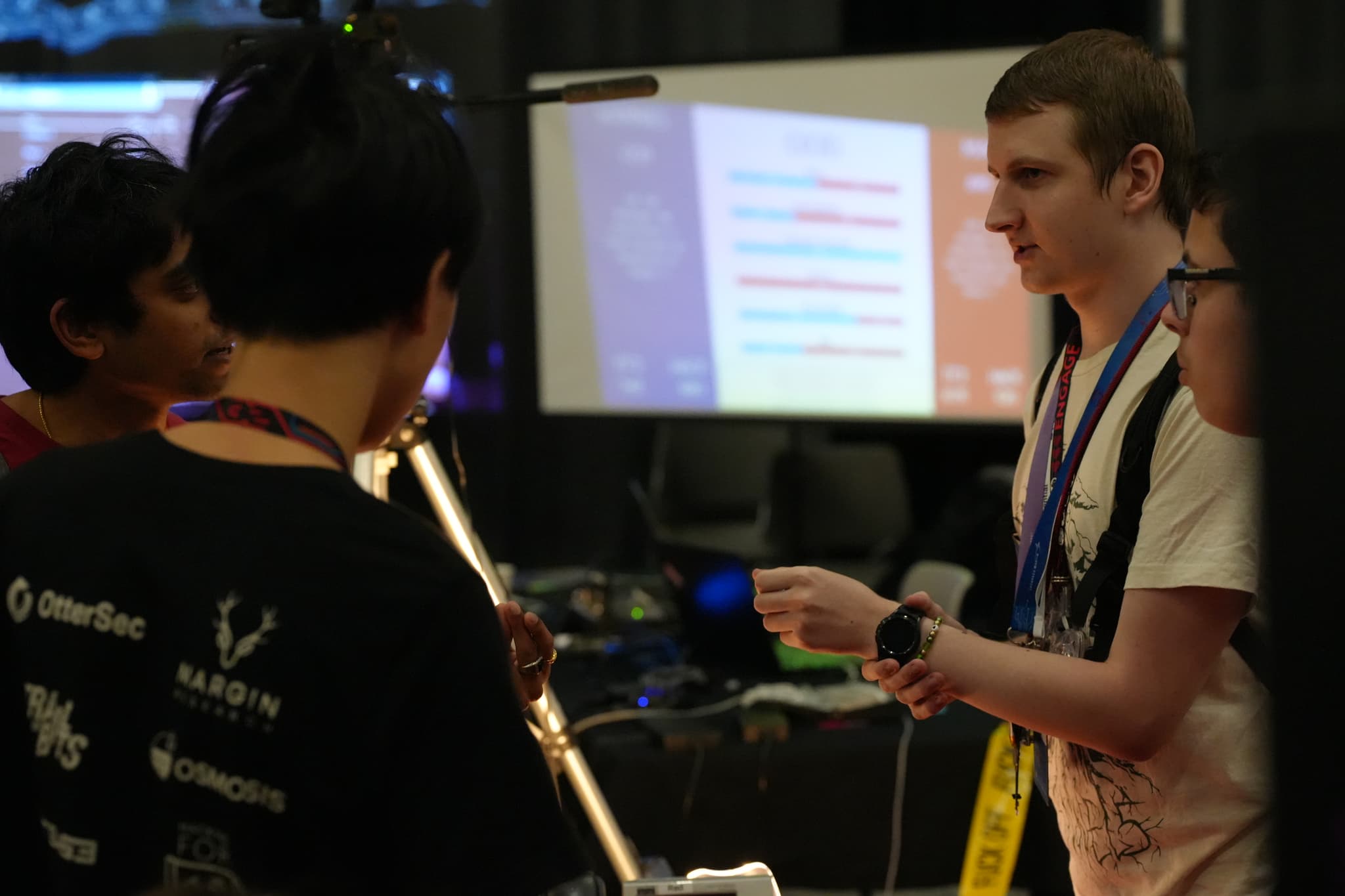 A group of people engaged in conversation at an indoor event, with one person holding a drink. A projector screen displaying text is visible in the background