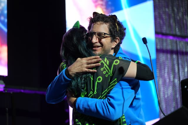 Two people embracing on a stage, one wearing a blue jacket and glasses, the other with dark hair and a black outfit with green patterns