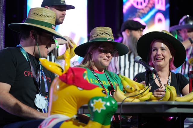 A group of people wearing straw hats and lanyards, sitting at a table with bananas and an inflatable kangaroo, with a colorful background and a referee in the back