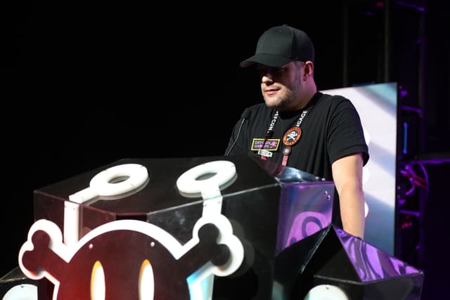 A person wearing a black cap and black shirt stands behind a podium with a cartoonish skull and crossbones design, illuminated by stage lights