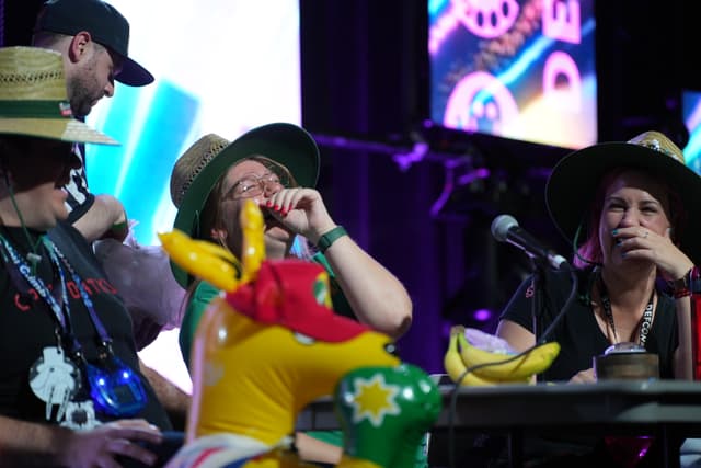 A group of people wearing hats are gathered around a table with microphones and various items, including an inflatable toy and bananas, in a lively setting with colorful lights in the background