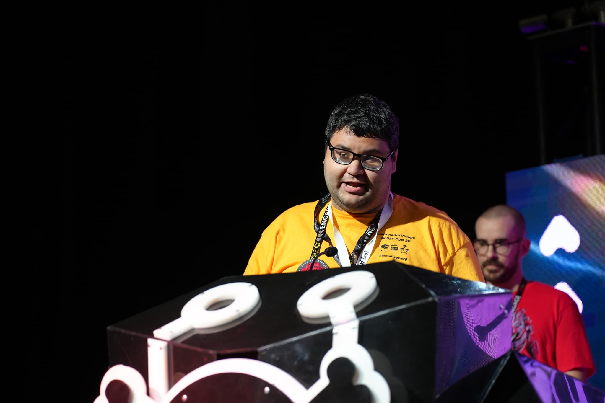 A person wearing a yellow shirt and glasses stands behind a podium with a stylized design, while another person in a red shirt stands slightly behind them. The background is dark with some colorful lighting