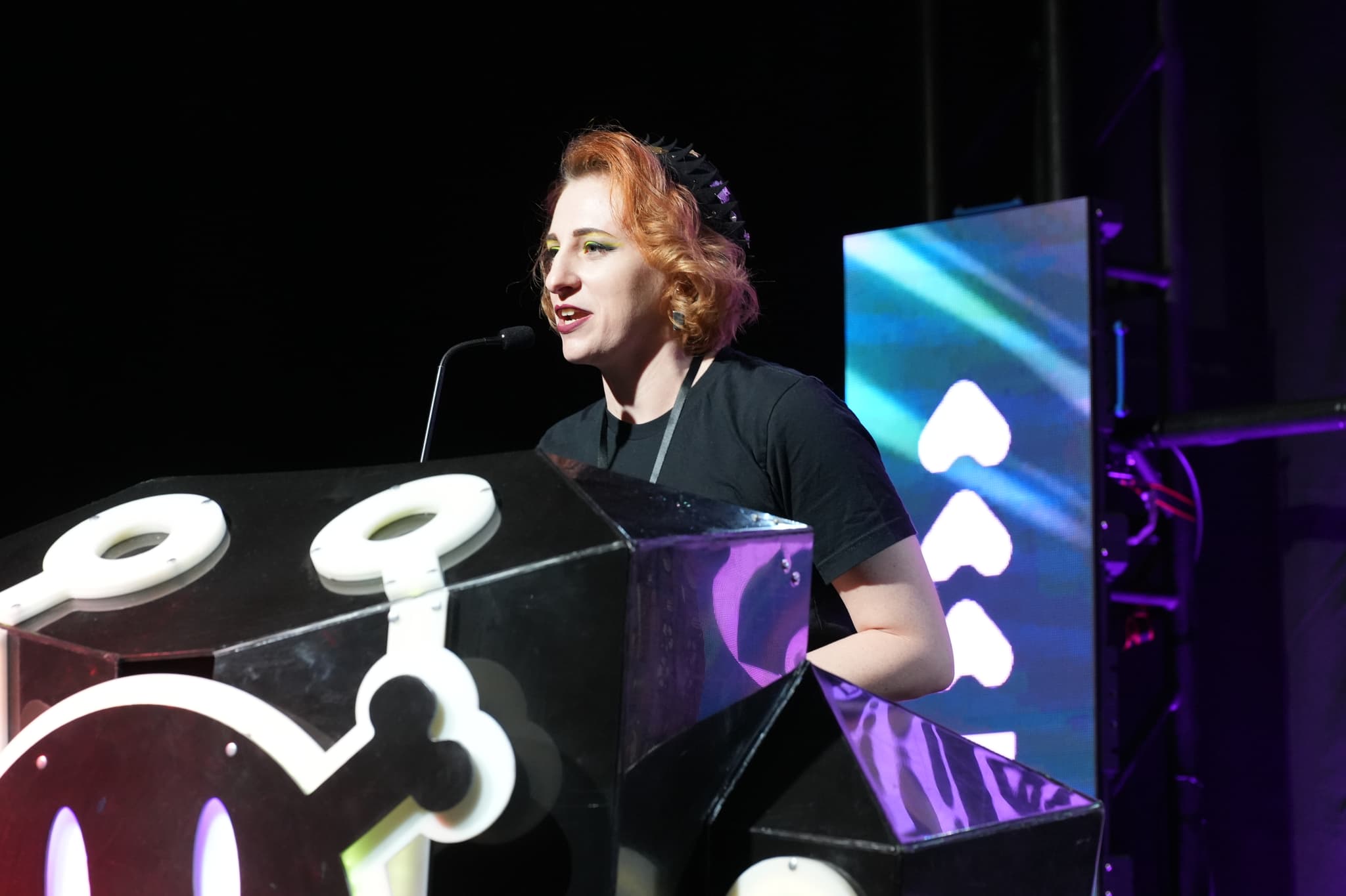 A person with short, reddish hair speaking at a podium decorated with a stylized design, with a microphone in front of them and a colorful, illuminated background