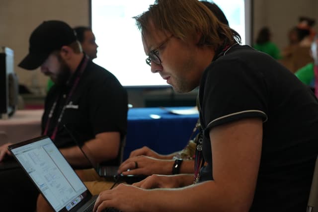 Two individuals working on laptops in a dimly lit room, with a large screen in the background