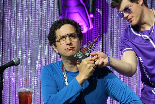 Two men are on stage with a sparkly curtain backdrop. One man in a blue shirt is holding a toy ray gun, while the other man in a purple shirt is pointing at it. A microphone and a drink are visible on the table in front of them