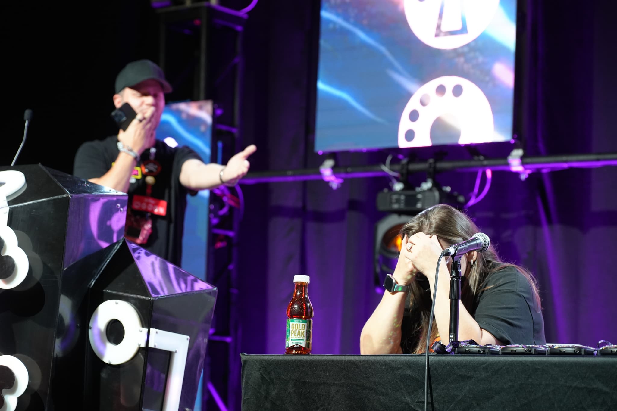 A person standing at a podium speaking into a microphone, while another person sits at a table with their head in their hands, appearing distressed. The background features a large screen and stage lighting