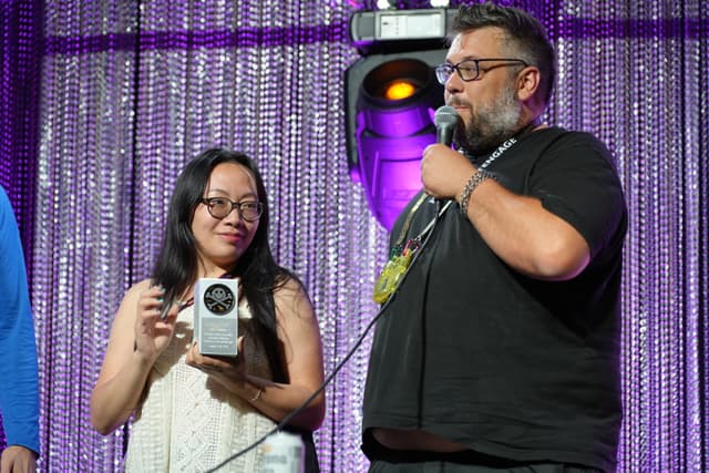 Two people on stage, one holding a microphone and the other holding a trophy, with a glittery curtain backdrop and stage lighting