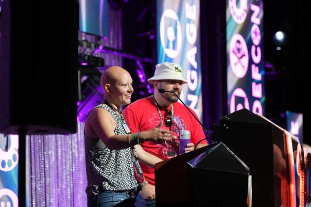 Two individuals standing at a podium, speaking into microphones, with a backdrop featuring the word DEFCON and various logos