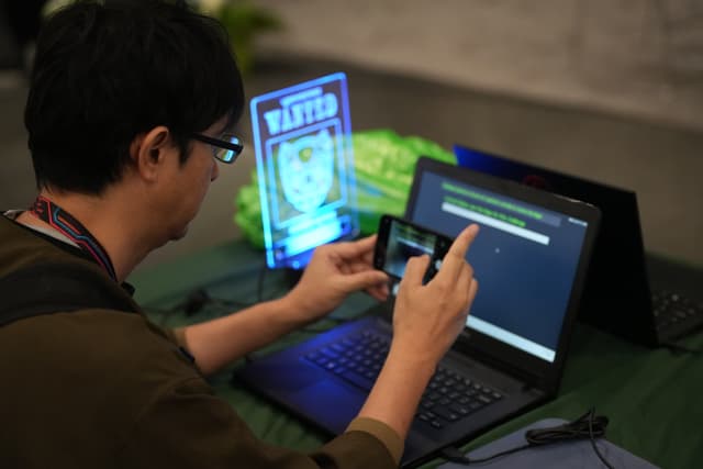 A person using a smartphone and a laptop simultaneously, with a WANTED poster displayed on a screen in the background