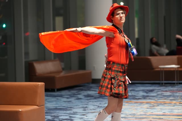 A person dressed in a vibrant orange outfit with a matching cape and hat, standing in a modern indoor setting with brown couches and large windows