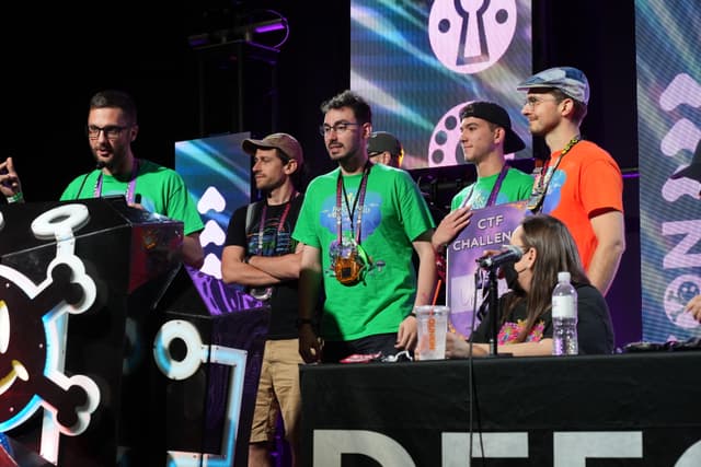 A group of people standing on a stage, some wearing medals, with a woman seated at a table with a microphone and water bottle. The background features colorful graphics and the word DEFCON