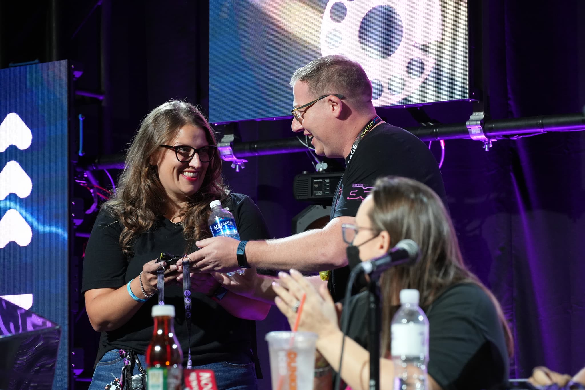 Three people on a stage, with one man handing a bottle of water to a smiling woman while another woman claps. The background features stage lighting and a large screen