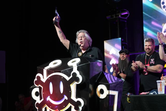 A person standing behind a podium with a skull and crossbones design, raising an object in the air, while others stand nearby clapping