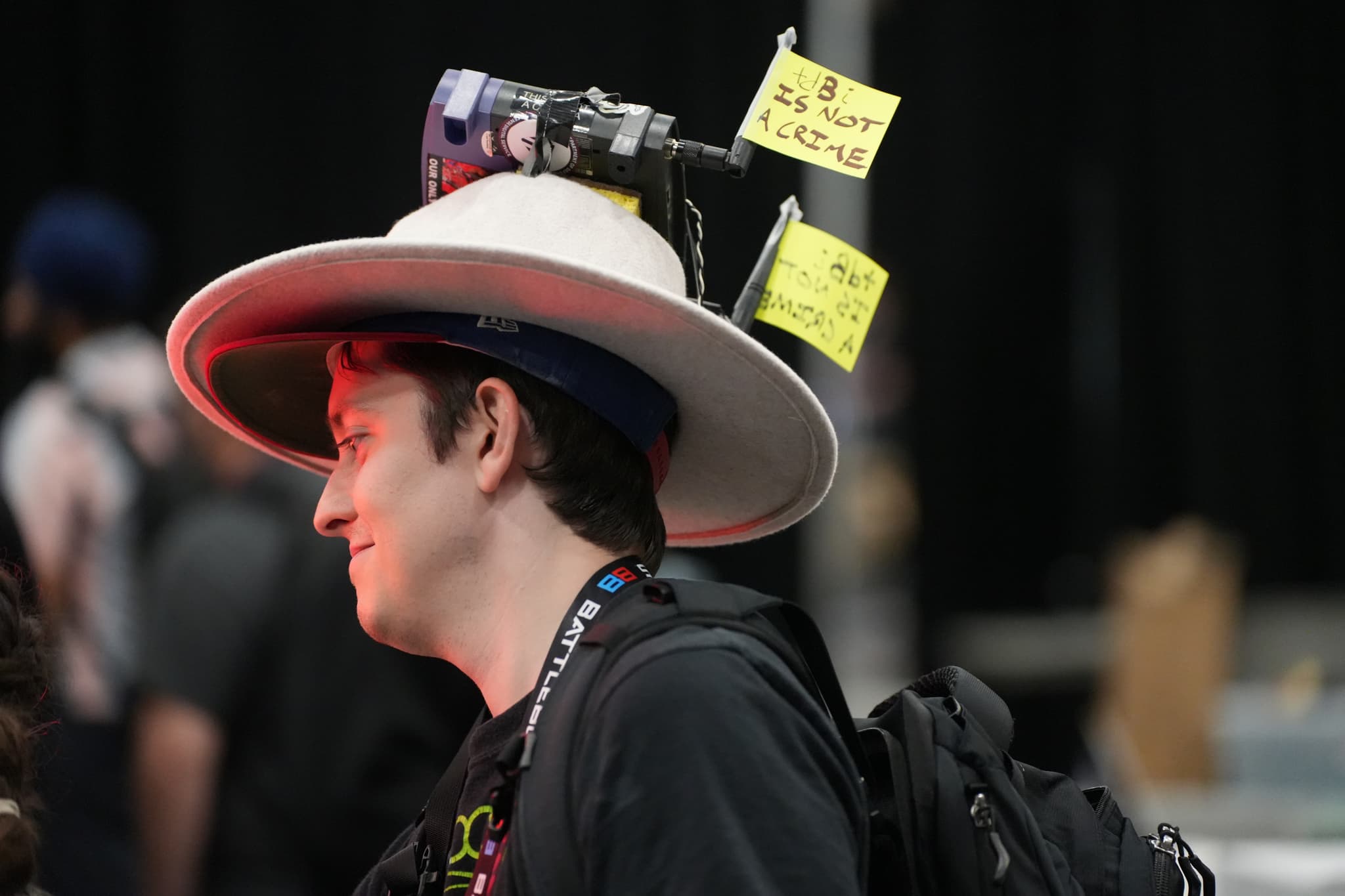 A person wearing a large hat with a camera and sticky notes attached to it, standing in a crowded indoor setting