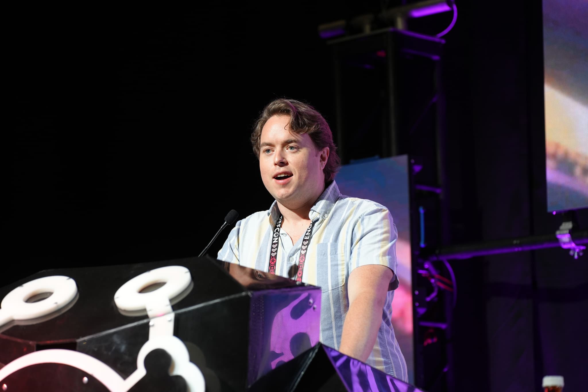 A person speaking at a podium with a microphone, with stage lighting and equipment in the background