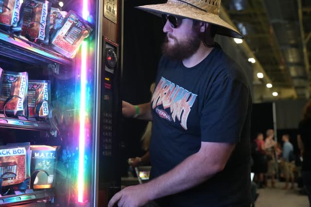 A man wearing a straw hat, sunglasses, and a graphic t-shirt is using a vending machine with colorful lights