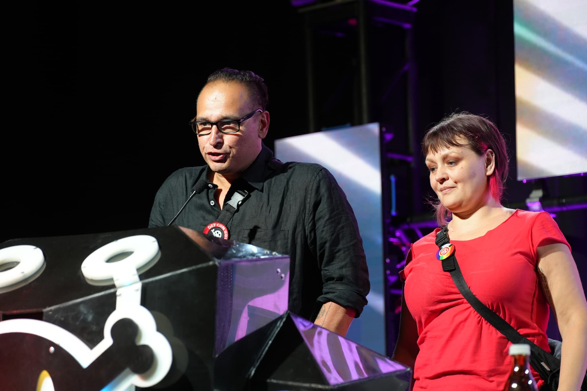 Two people standing on a stage, one speaking at a podium with a microphone, and the other standing beside them