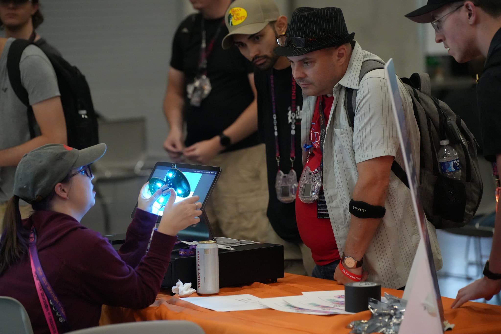 A group of people gathered around a table, with one person seated and holding up a device displaying an image, while others stand and observe