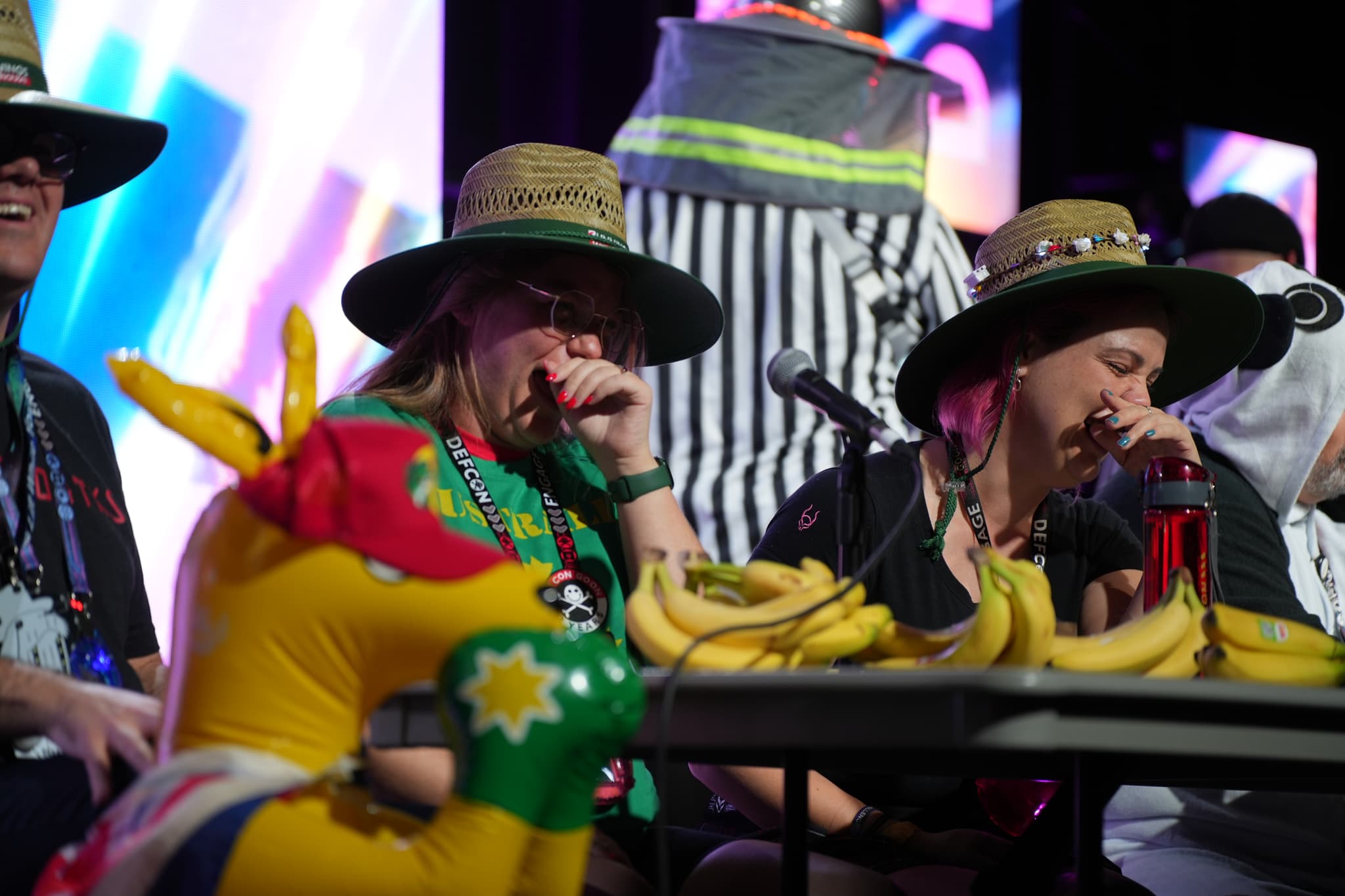 Several people wearing hats are seated at a table with a large number of bananas and a colorful inflatable animal. The background features bright, colorful lights