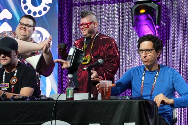 Four people at a panel event, with one person in a blue shirt seated at a table, and three others standing behind. The background features a purple curtain and stage lighting