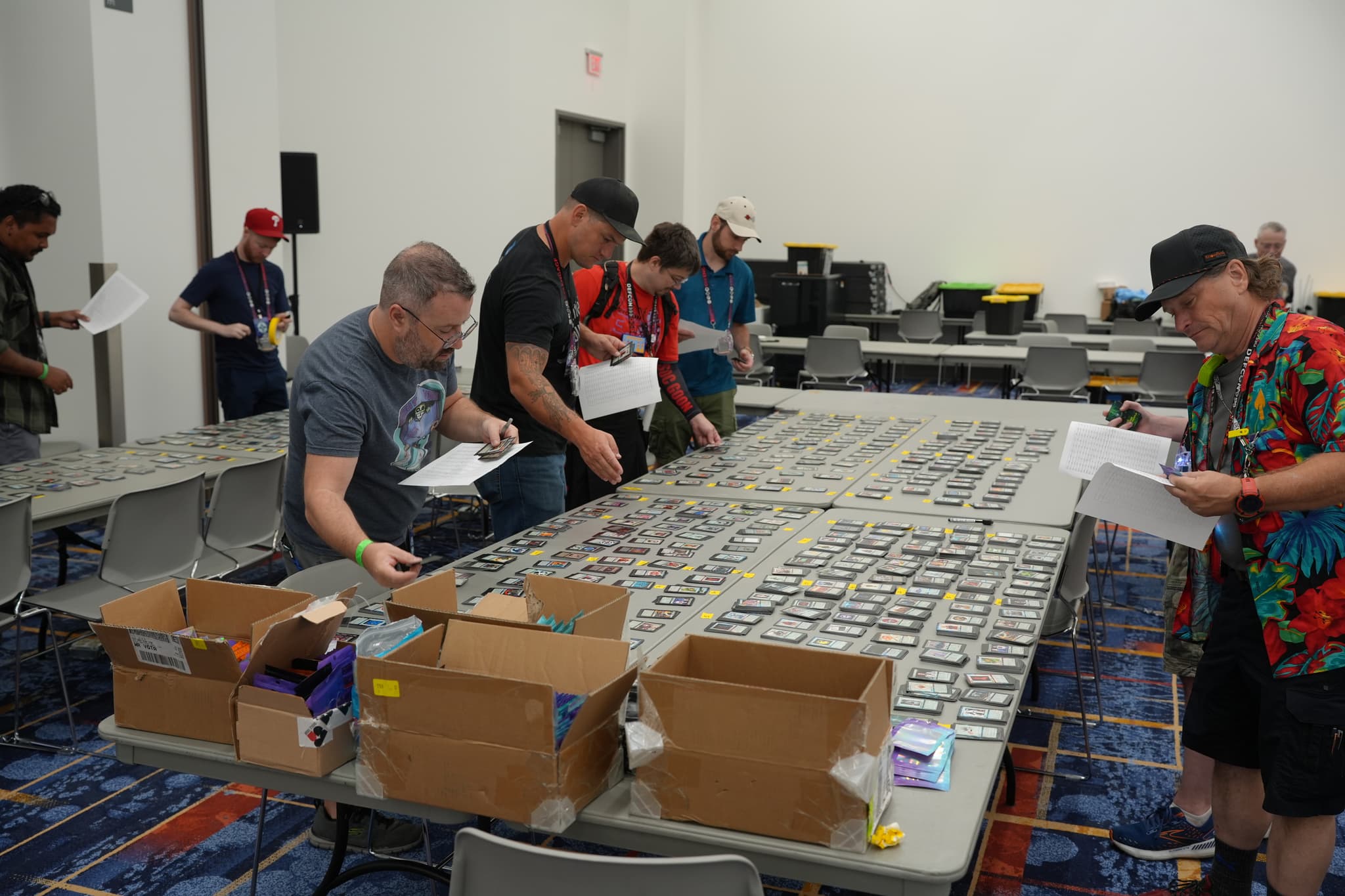 Several people examining and sorting through a large collection of trading cards spread out on tables in a room, with boxes and papers around them