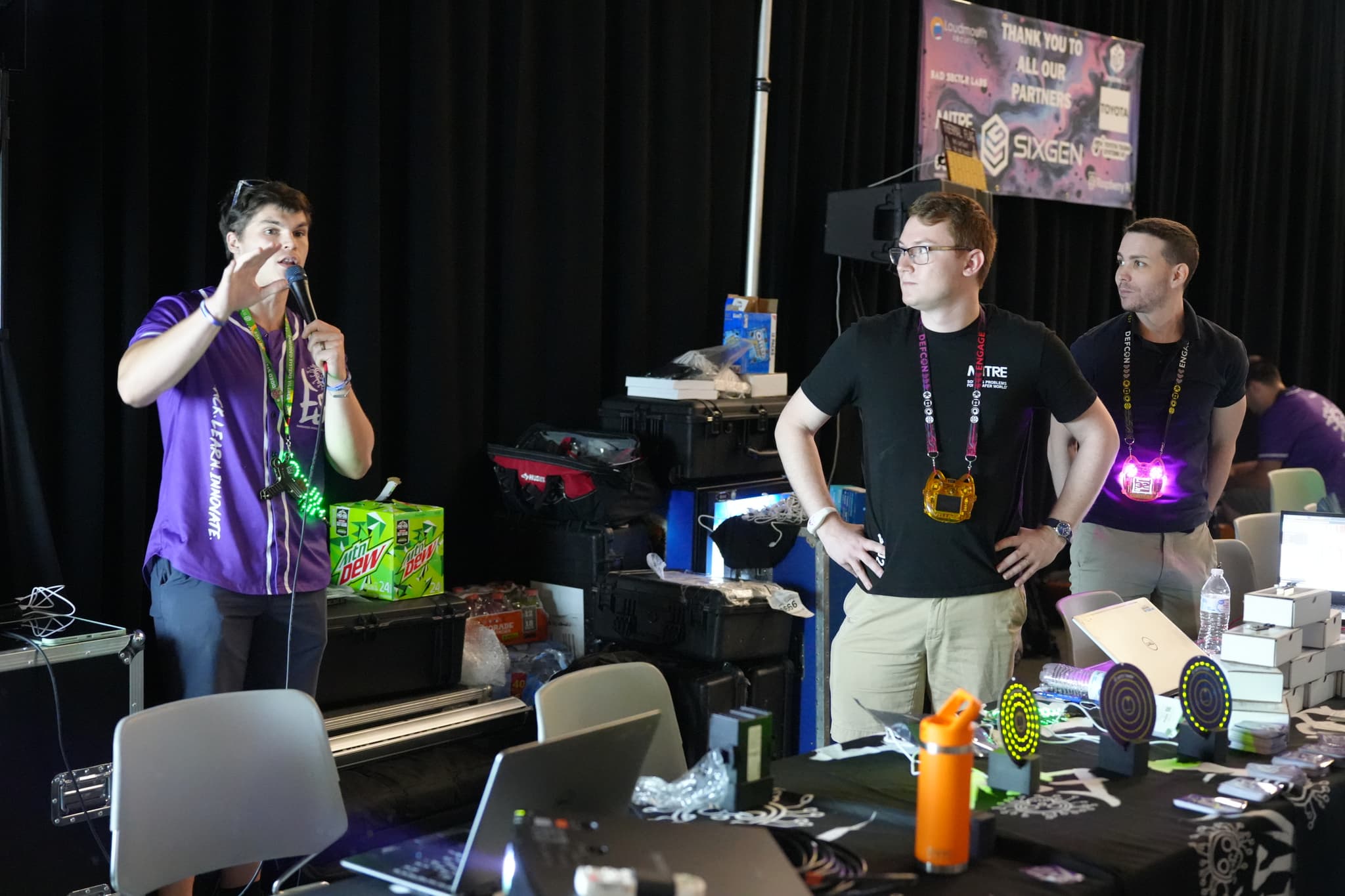 Three people standing at a cluttered table with laptops and various items, engaged in conversation. One person is gesturing with their hands, while the other two are looking in the same direction. The background includes a banner and various equipment