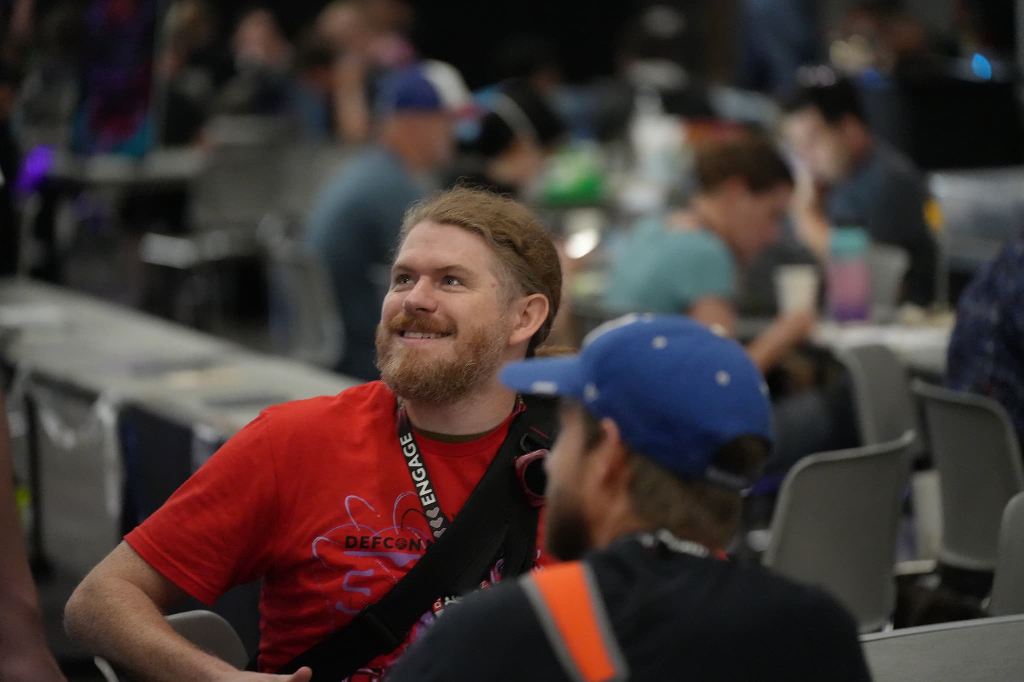 A bearded man in a red shirt is smiling and looking to the side while sitting in a crowded indoor setting with people engaged in various activities in the background