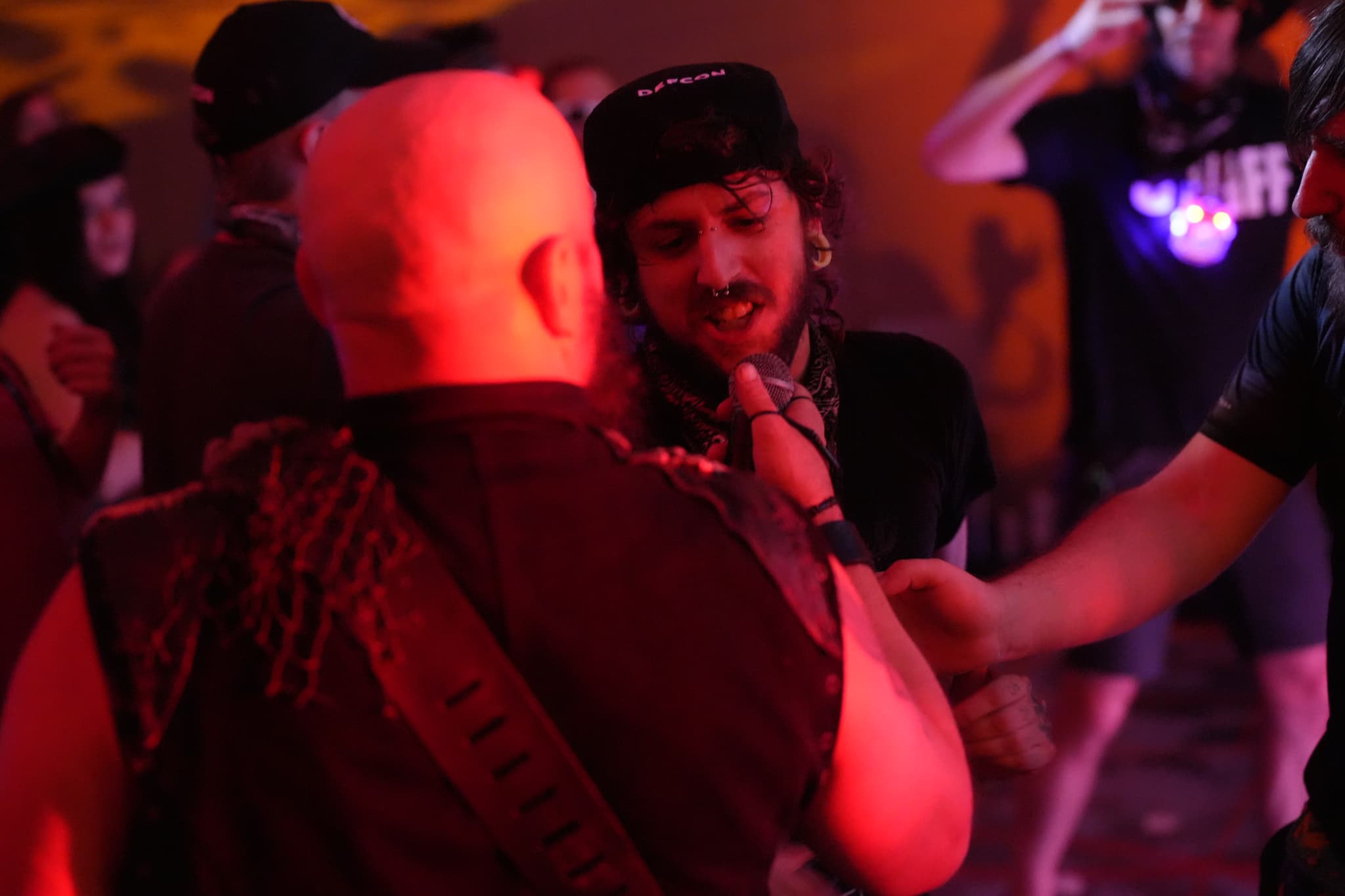 A group of people in a dimly lit, red-hued environment, with one person in the foreground facing away from the camera and another person facing them, appearing to be engaged in conversation or interaction