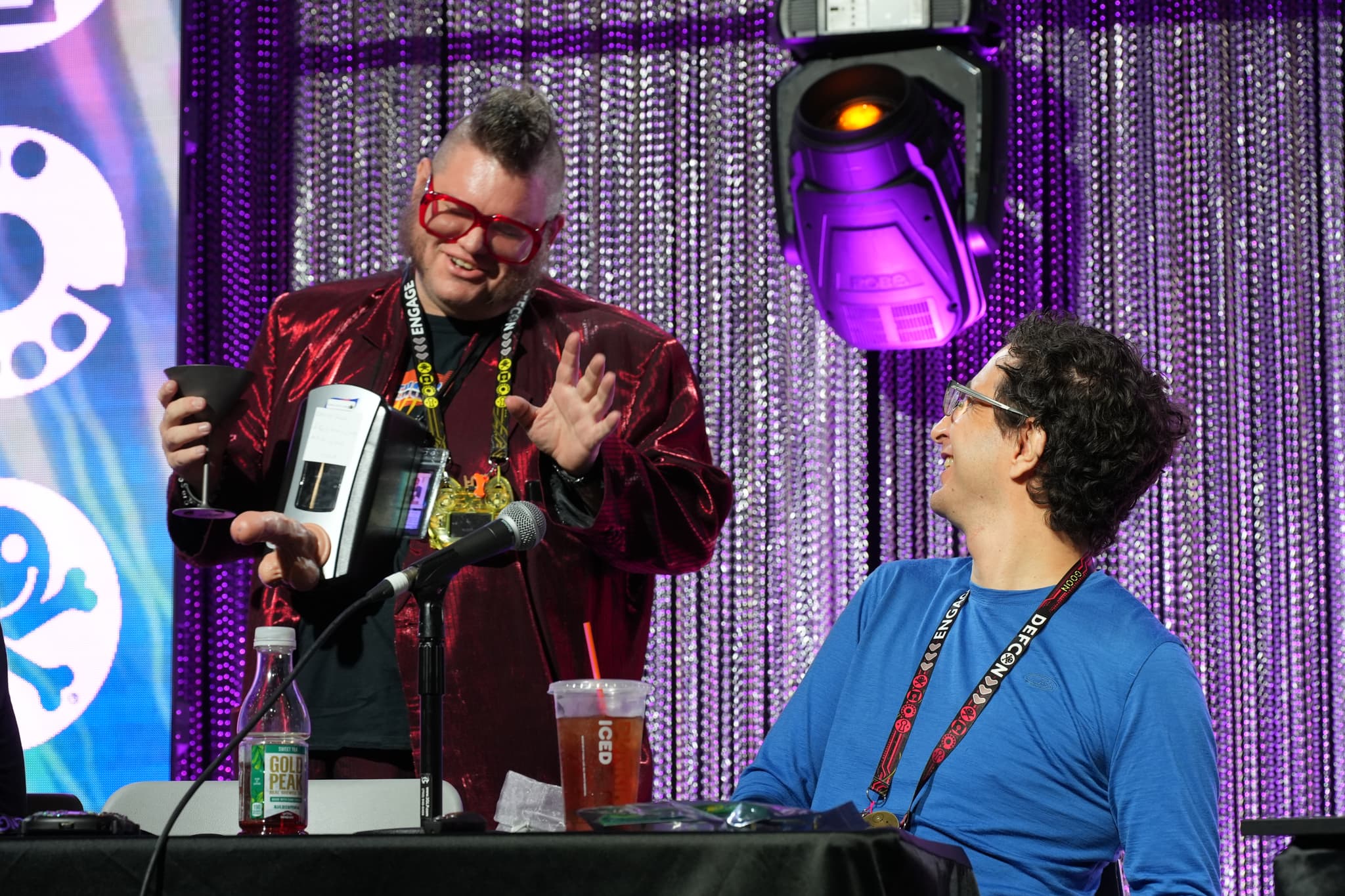 Two individuals at a conference or event, one standing and holding a device while wearing red glasses and a shiny jacket, and the other sitting and smiling, wearing a blue shirt. They are in front of a table with microphones and drinks, with a purple curtain backdrop