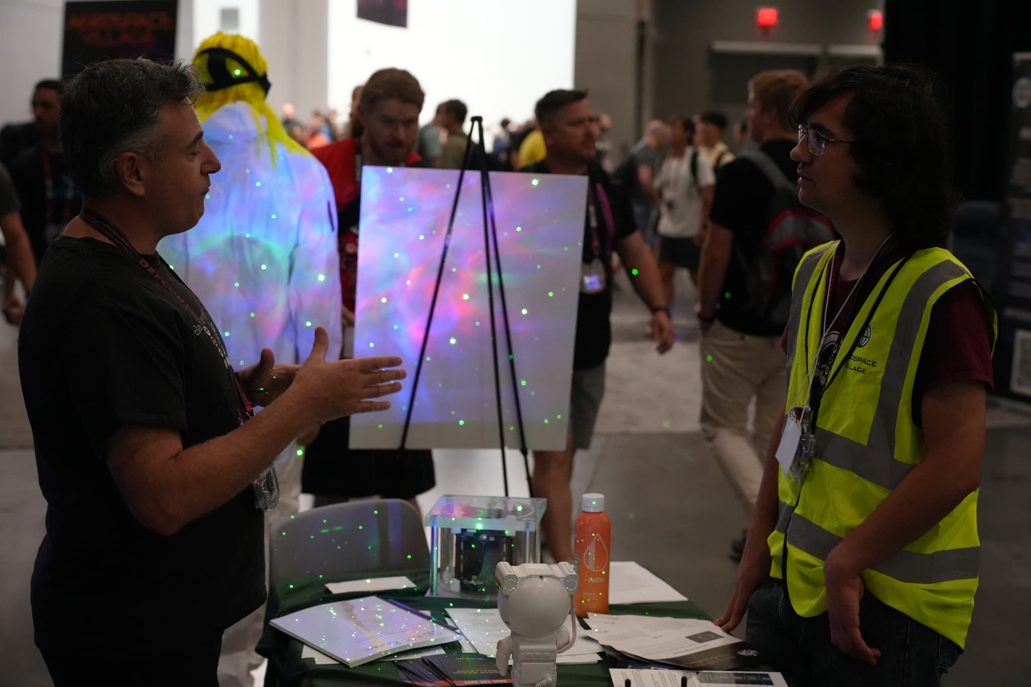 Two people engaged in conversation at a booth in a busy convention or exhibition hall, with one person wearing a high-visibility vest and the other gesturing with their hands. A colorful, illuminated display is visible in the background