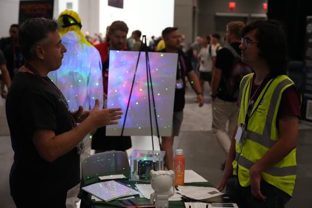 Two people engaged in conversation at a booth in a busy convention or exhibition hall, with one person wearing a high-visibility vest and the other gesturing with their hands. A colorful, illuminated display is visible in the background